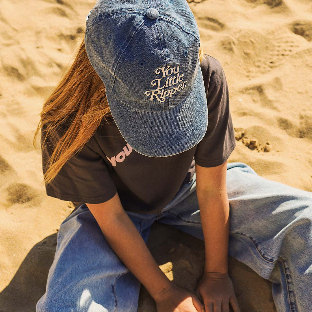Kid sitting on the beach wearing the You Little Ripper cotton cap in blue