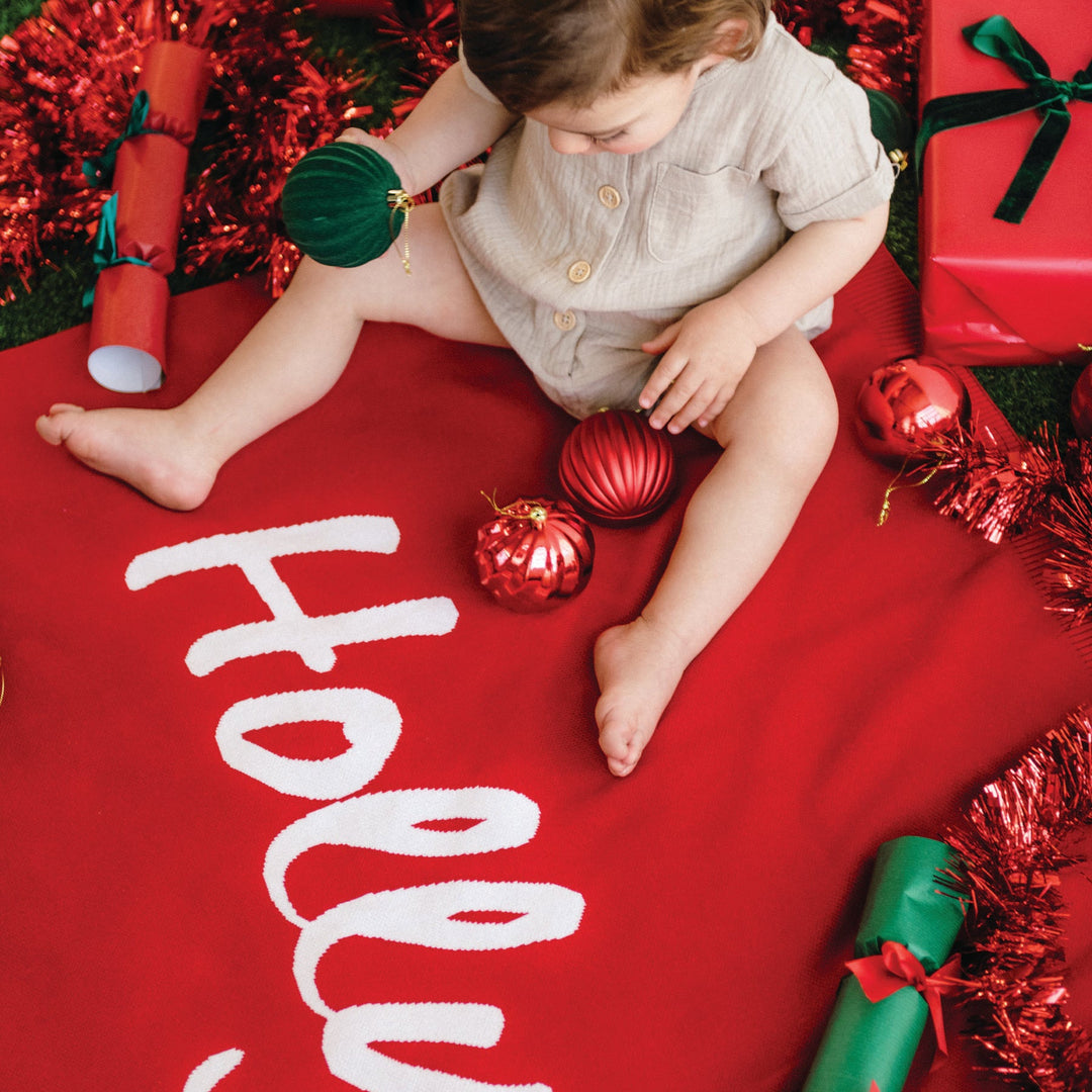 Child sitting on a red blanket with Christmas decorations and the word &