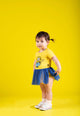 A baby girl in a Bananas Minion Tulle Bodysuit by Rock Your Baby Online standing in front of a yellow background.