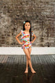 A little girl in a Rock your Baby Online BUTTERFLIES ONE-PIECE SWIM swimsuit posing in front of a brick wall.